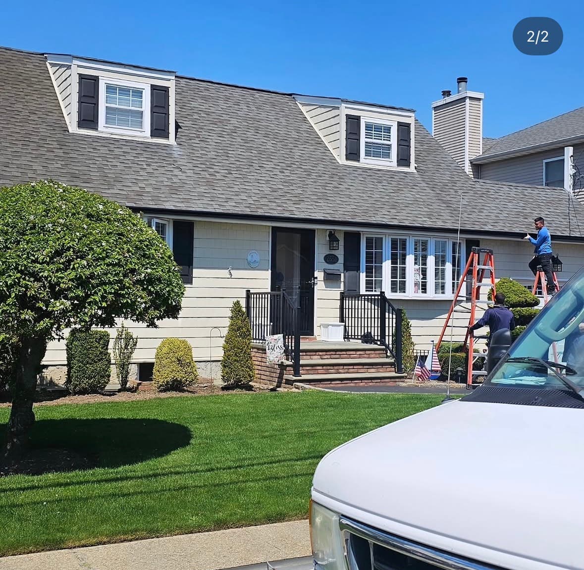 Crew on ladders working along the eaves of a cream-shingled Cape with white dormers on a clear day.