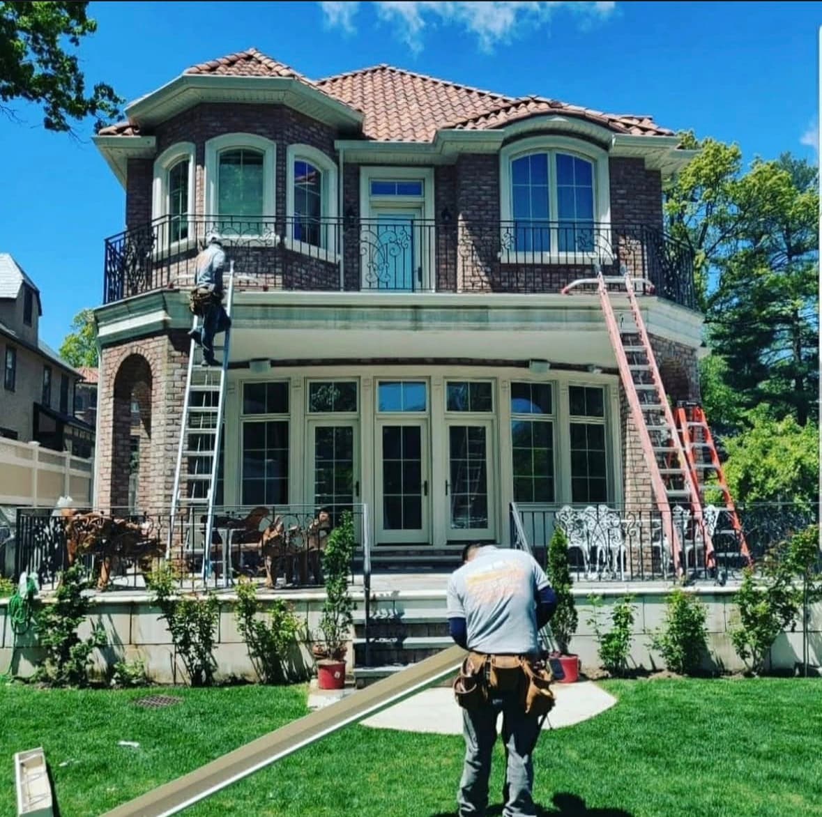 A crew member carrying a single continuous length of seamless gutter across the lawn of a large brick home, with ladders set against the second story.